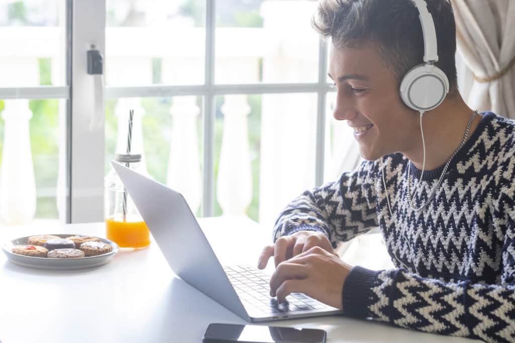 Handsome, smiling teenager using laptop at home in front of the window wearing headphones
